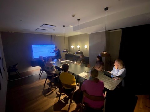 Kuvassa näkyy ryhmä ihmisiä himmeästi valaistussa kokoushuoneessa. Huoneessa on suuri näyttö yhdellä seinällä ja pitkä pöytä tuoleineen. Ihmiset istuvat pöydän ympärillä ja vaikuttavat olevan esityksen tai keskustelun parissa.